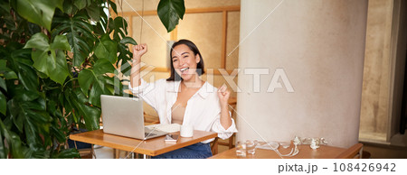 Happy young asian woman winning on laptop, receive good news, achieve goal at work, triumphing and smiling pleased, sitting in cafe 108426942