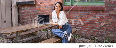 Vertical shot of good-looking asian woman with laptop, sitting in outdoor cafe, drinking coffee and smiling, laughing carefree Vertical shot of good-looking asian woman with laptop, sitting in outdoor cafe, drinking coffee and smiling, laughing carefree 108426984