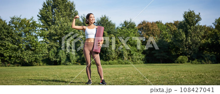 Portrait of fit and healthy young fitness woman, shows her biceps with pleased smiling face, stands with yoga rubber mat on grass in park 108427041