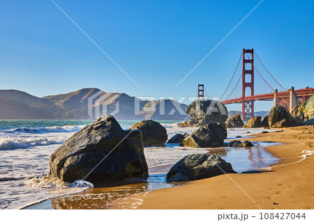 Sunny beach with waves crashing against boulders and distant Golden Gate Bridge 108427044