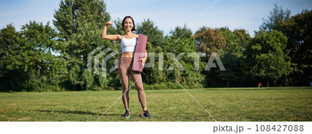Confident and sporty young asian woman, standing in park with rubber mat, wearing sportswoman, smiling at camera, workout on fresh air Confident and sporty young asian woman, standing in park with rubber mat, wearing sportswoman, smiling at camera, workout on fresh air 108427088