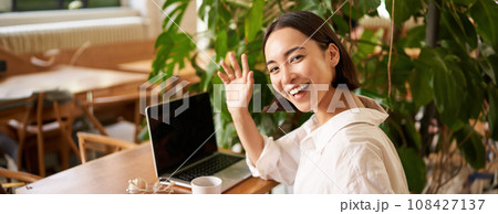 Portrait of beautiful young woman wave at you, say hello, greeting friend while sitting in cafe with laptop and waiting for someone Portrait of beautiful young woman wave at you, say hello, greeting friend while sitting in cafe with laptop and waiting for someone 108427137