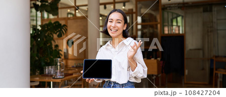 Friendly smiling asian woman, cafe manager, showing tablet screen and looking happy, demonstrating her restaurant profit Friendly smiling asian woman, cafe manager, showing tablet screen and looking happy, demonstrating her restaurant profit 108427204
