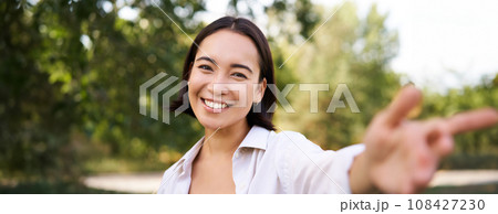 People and lifestyle. Happy asian woman takes selfie in park, photo on smartphone, smiling and looking joyful People and lifestyle. Happy asian woman takes selfie in park, photo on smartphone, smiling and looking joyful 108427230