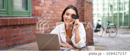 Happy young asian female, sitting with laptop and showing credit card, paying bills online, shopping in internet, sitting in cafe with coffee 108427237