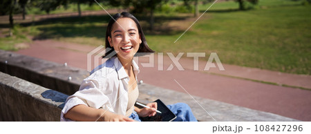 Happy korean woman sitting on bench with digital tablet and graphic pen, smiling, turning back at camera and talking lively Happy korean woman sitting on bench with digital tablet and graphic pen, smiling, turning back at camera and talking lively 108427296