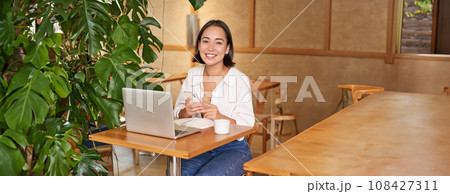 Vertical shot of young asian girl sits in cafe with laptop and smartphone, relaxing and surfing the net, working remotely 108427311