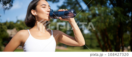 Fit asian sportswoman drinks water while running marathon. Fitness girl jogging in park, staying hydrated. Fit asian sportswoman drinks water while running marathon. Fitness girl jogging in park, staying hydrated. 108427320