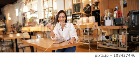 Young stylish asian woman, business owner in glasses, sitting in cafe with notebook, smiling at camera Young stylish asian woman, business owner in glasses, sitting in cafe with notebook, smiling at camera 108427446