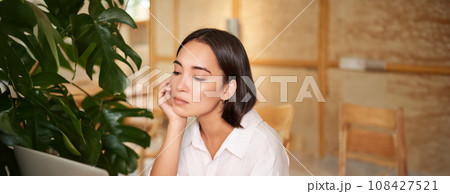 Vertical shot of asian girl looking tired at laptop screen, bored of working, sitting in cafe sleepy and exhausted 108427521