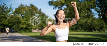 Happy fitness girl achieve goal, finish marathon, running with hands up, celebrating victory while jogging, triumphing in park 108427553