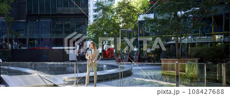 Corporate woman in suit, standing with confidence in beige suit with working documents outdoors, posing in city center Corporate woman in suit, standing with confidence in beige suit with working documents outdoors, posing in city center 108427688