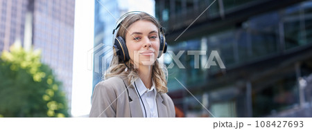 Portrait of smiling corporate woman, girl in suit listening music in headphones, sitting in city centre 108427693
