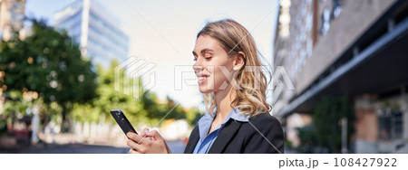Corporate woman stands on street and texts message on mobile phone, smiling while looking at smartphone screen Corporate woman stands on street and texts message on mobile phone, smiling while looking at smartphone screen 108427922