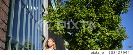Portrait of confident saleswoman in her 30s, posing near office building on street, wearing beige suit, holding folder with documents Portrait of confident saleswoman in her 30s, posing near office building on street, wearing beige suit, holding folder with documents 108427944