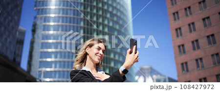Portrait of businesswoman wave her hand at mobile phone camera, waves hand during video chat, stands in suit in city center outdoors 108427972