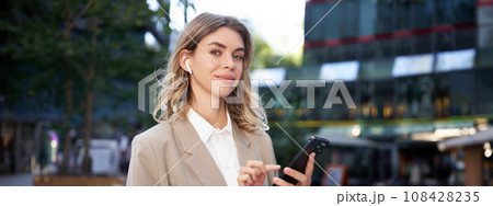 Smiling beautiful corporate woman in beige suit, standing on street in city with wireless headphones, holding smartphone 108428235