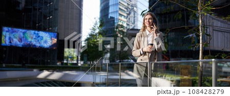 Corporate woman stands on street in city center, has a phone call, telephone conversation, smiling and drinking coffee 108428279