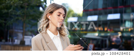 Successful businesswoman in beige suit, wireless headphones, looking at mobile phone, using smartphone app, standing outdoors on street Successful businesswoman in beige suit, wireless headphones, looking at mobile phone, using smartphone app, standing outdoors on street 108428401