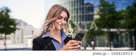 Close up portrait of young corporate woman in black suit, holds smartphone, texting message while standing on street, using mobile application 108428423