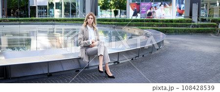 Businesswoman sitting alone on empty street near fountain. Female entrepreneur outdoors in suit and heels 108428539