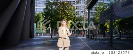 Beautiful businesswoman in beige suit, standing near office buildings on street, posing with confident, relaxed face expression Beautiful businesswoman in beige suit, standing near office buildings on street, posing with confident, relaxed face expression 108428578