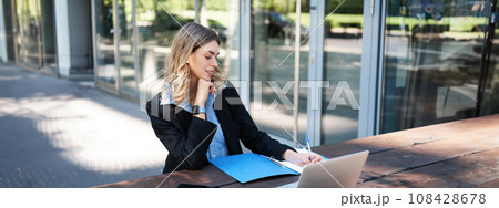 Successful businesswoman working outdoors. Corporate woman sitting on bench with laptop, writing, taking notes during work meeting 108428678