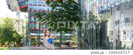 Portrait of young saleswoman in beige business suit, holding blue folder with work documents, standing outdoors on street of city center 108428717