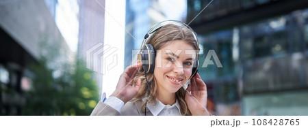 Happy woman listens to music in headphones while walking in city centre, smiling Happy woman listens to music in headphones while walking in city centre, smiling 108428765