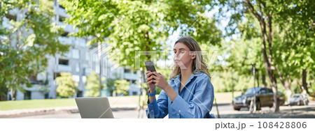 Portrait of businesswoman working outside in park, sitting with laptop, work documents and using mobile phone, text message, reading on smartphone Portrait of businesswoman working outside in park, sitting with laptop, work documents and using mobile phone, text message, reading on smartphone 108428806