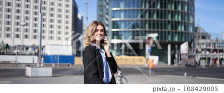 Portrait of businesswoman in suit, standing on street with mobile phone, on call with someone, smiling while having telephone conversation Portrait of businesswoman in suit, standing on street with mobile phone, on call with someone, smiling while having telephone conversation 108429005