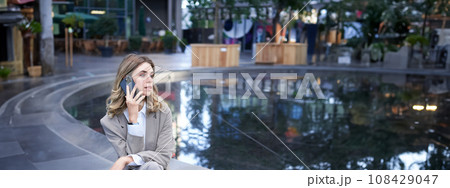 Vertical shot of corporate woman sitting outdoors with laptop, talking on mobile phone, working ouside office building while waiting for someone Vertical shot of corporate woman sitting outdoors with laptop, talking on mobile phone, working ouside office building while waiting for someone 108429047