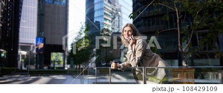 Happy smiling saleswoman stands on street on coffee break, talking on mobile phone and drinks takeaway drink Happy smiling saleswoman stands on street on coffee break, talking on mobile phone and drinks takeaway drink 108429109