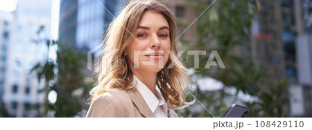 Close up portrait of corporate woman, young intern walks on street to office, holds mobile phone, texts messages on smartphone Close up portrait of corporate woman, young intern walks on street to office, holds mobile phone, texts messages on smartphone 108429110