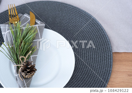 Serving a festive table with plates, forks, knives, napkins close-up tissue in a cage decorated for Christmas and New Year pine branch with a cone. Eco-style, minimalism 108429122