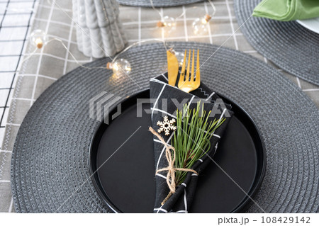 Serving a festive table with plates, forks, knives, napkins close-up tissue in a cage decorated for Christmas and New Year pine branch with a cone. Eco-style, minimalism 108429142