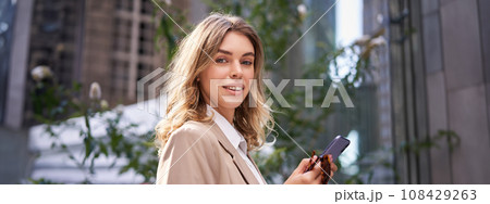 Close up portrait of smiling businesswoman, young office intern in suit, holding smartphone and looking happy at camera, standing on street 108429263