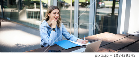 Happy saleswoman taking notes, working outdoors on fresh air, sitting with laptop outside office. Corporate woman laughing, writing on piece of paper Happy saleswoman taking notes, working outdoors on fresh air, sitting with laptop outside office. Corporate woman laughing, writing on piece of paper 108429345