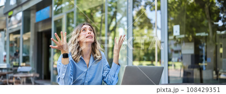 Disappointed young businesswoman feeling frustrated, sitting with laptop outdoors on street, looking up and complaining 108429351