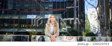 Young beautiful businesswoman standing in beige suit on sunny street near office buildings 108429703