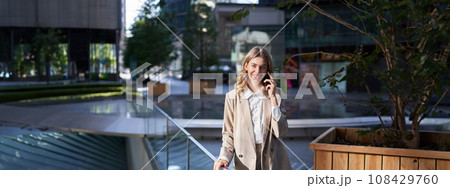 Successful businesswoman talking on mobile phone with confident smile, dealing with business calls, standing on street in city 108429760