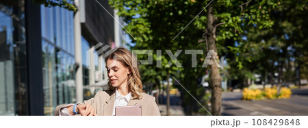Vertical portrait of beautiful businesswoman in suit, holds her digital tablet, looks at watch on wrist, reads message or checks time 108429848