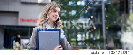 Smiling professional businesswoman talking on mobile phone, holding laptop and work folders in hand, having a business call Smiling professional businesswoman talking on mobile phone, holding laptop and work folders in hand, having a business call 108429894