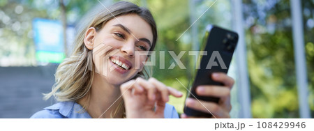 Beautiful young woman sitting outdoors on street, smiling at smartphone, buying online, order on mobile phone Beautiful young woman sitting outdoors on street, smiling at smartphone, buying online, order on mobile phone 108429946