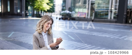 Businesswoman sitting with laptop, wearing suit, writing down notes in her planner, being in city centre outdoors Businesswoman sitting with laptop, wearing suit, writing down notes in her planner, being in city centre outdoors 108429966
