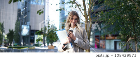Smiling businesswoman standing on street, holding work documents, laptop and drinking takeaway coffee 108429973