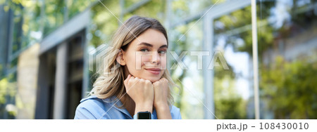 Close up portrait of smiling blond woman, student with digital watch, looking happy at camera, sitting outside on street 108430010