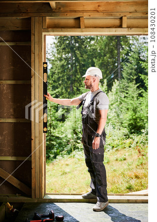 Carpenter building wooden-framed house near the forest. Bearded man inspects the walls for levelness using a spirit level. Concept of environmentally friendly construction. 108430201