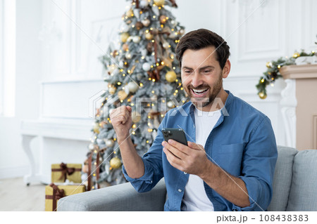 Man celebrating New Year and Christmas at home sitting on sofa, celebrating victory and successful news received on phone, using application on smartphone. 108438383