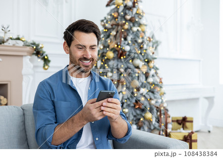 Man celebrating New Year and Christmas alone at home, using app on phone, sending party invitations, smiling, holding smartphone, sitting on sofa in living room. 108438384